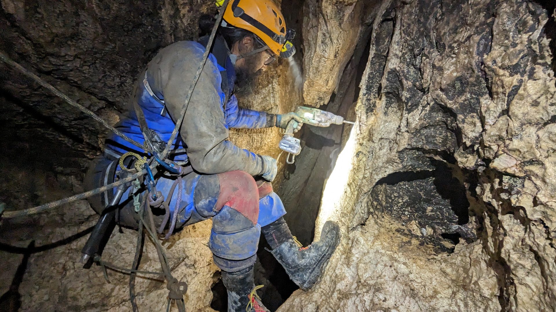 A caver drilling a hole at the top of a pitch while attached to a traverse line