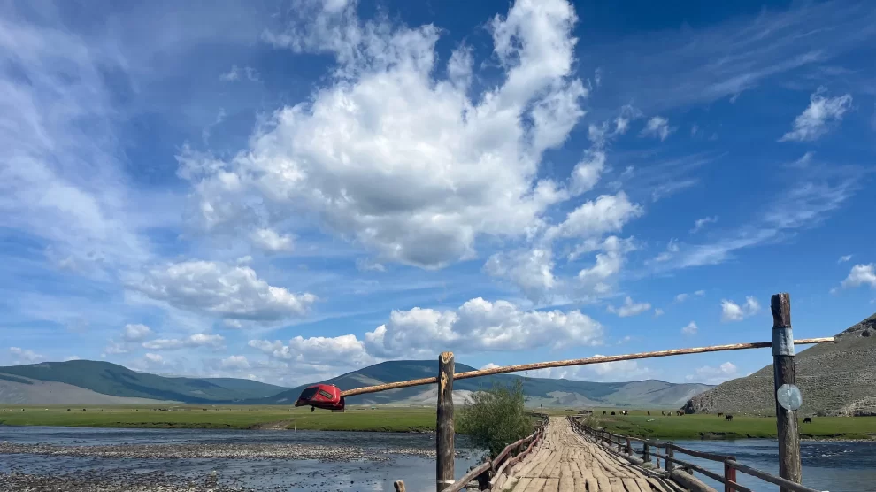 A wooden bridge leading away from the camera crosses a large river in the Darkhad depression. Behind the bridge a large flat plain extends into the distance where mountains can be seen.