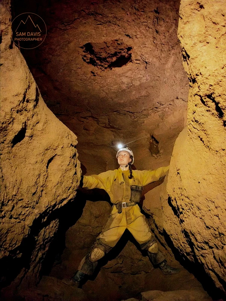 A female caver wearing a yellow caving suit straddles the cave passage.