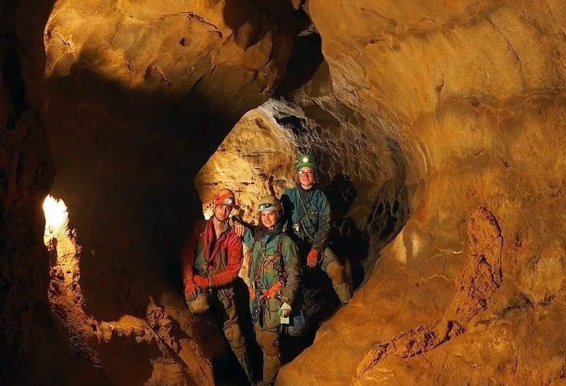 Three cavers smiling and facing the camera within a muddy cave passage