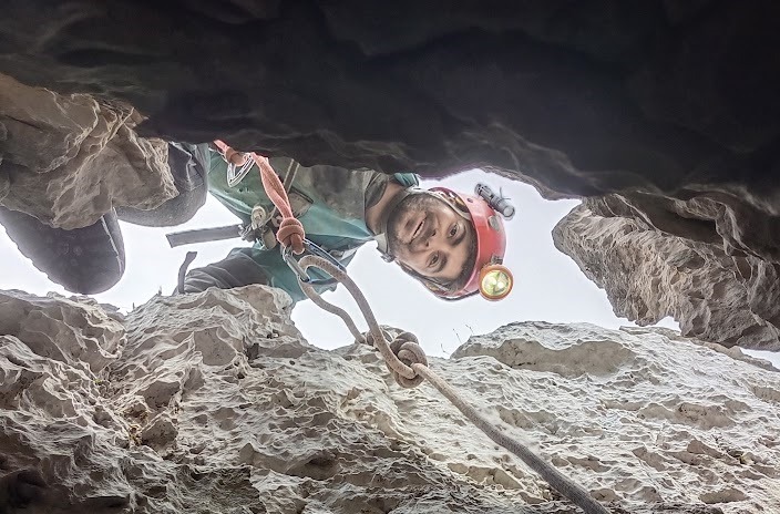 Arun looking down into a cave entrance while attached to a rope.